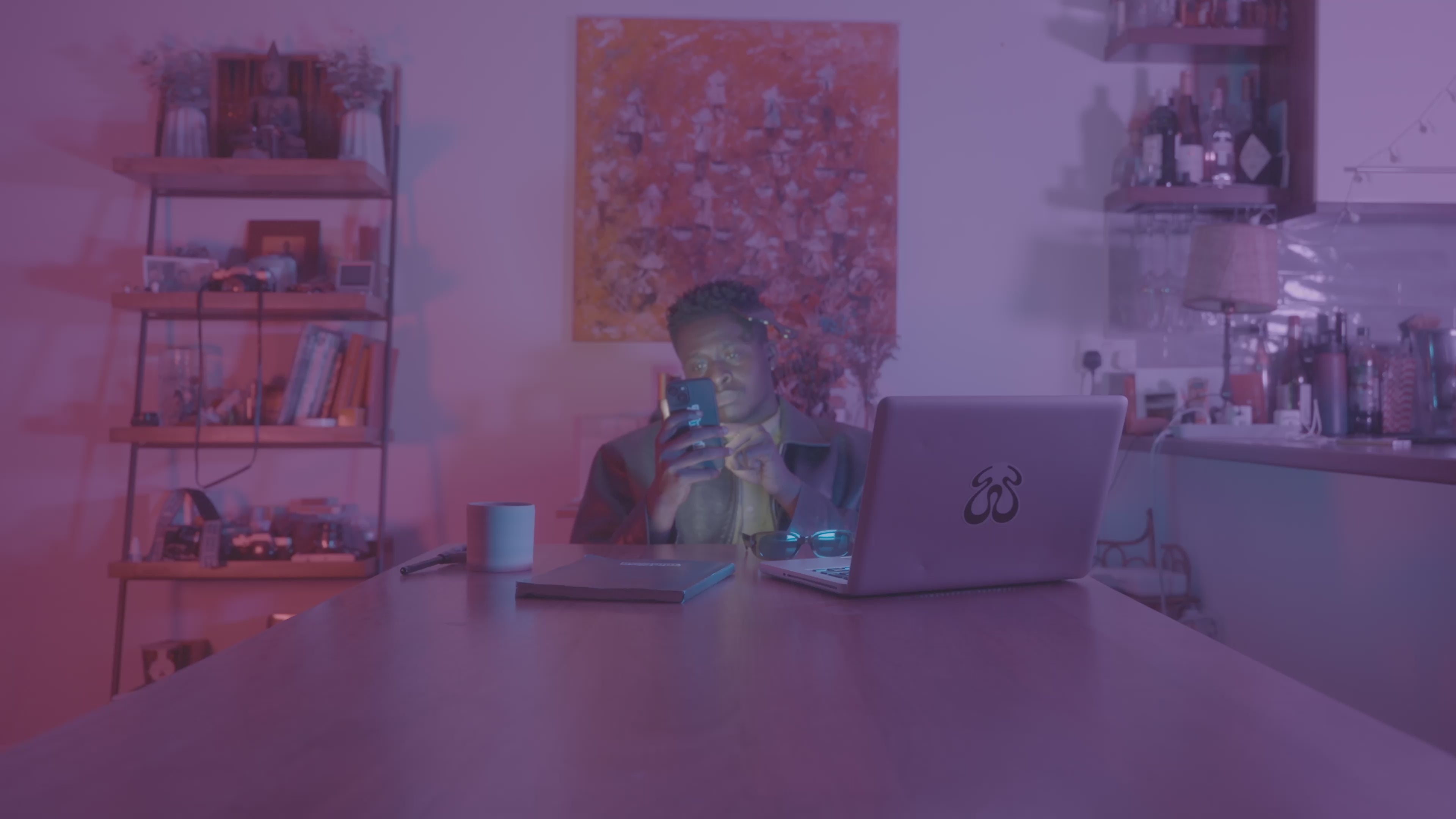 Man sitting at a desk, staring at his phone under purple light, surrounded by digital distractions.